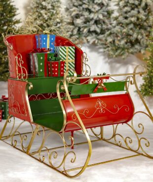 life-size classic two-seater elegant metal Christmas sleigh with a glossy red and green finish with metallic gold accents in front of mailboxes and trees and metal gift boxes on a seat