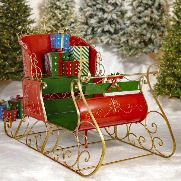 life-size classic two-seater elegant metal Christmas sleigh with a glossy red and green finish with metallic gold accents in front of mailboxes and trees and metal gift boxes on a seat