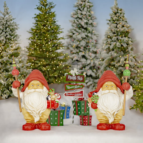 Two tall Santa gnome statues in red hats holding gifts and staffs in front of Christmas background
