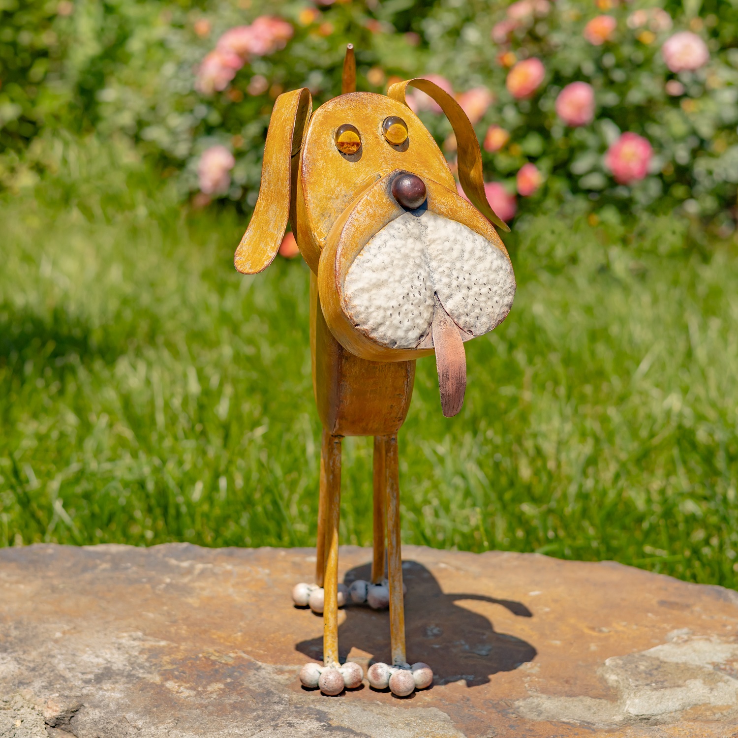 This is a whimsical 3D metal sculpture of a dog, about knee-high, with a round body, thin curved legs, and a tail raised in the air. Its large, textured snout is white, with a pink tongue playfully sticking out. The ears are long and floppy, and the eyes are small, raised circles. The sculpture has a warm, rusty brown hand-painted finish and stands on a flat stone surface in a sunny garden with green grass and blooming flowers behind it.