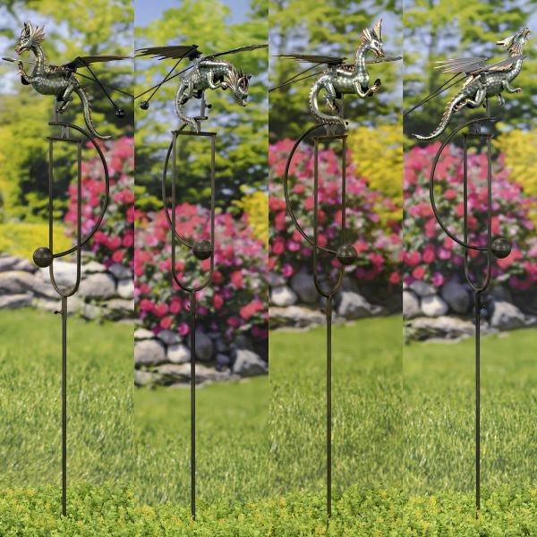This image show 4 metallic dragon sculpture perched on a garden stake. The dragon has large wings spread out, a long tail, and is balanced by thin rods with weights at the ends, allowing it to rock gently in the wind. The background is a blurred garden scene with green trees and colorful flowers.