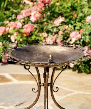 closeup of the basin with 3 small bird sculptures perched on the perimeter in a distressed antique copper-bronze finish - part of a metal birdbath with folding tristand standing on a rock floor surrounded by pink flowers