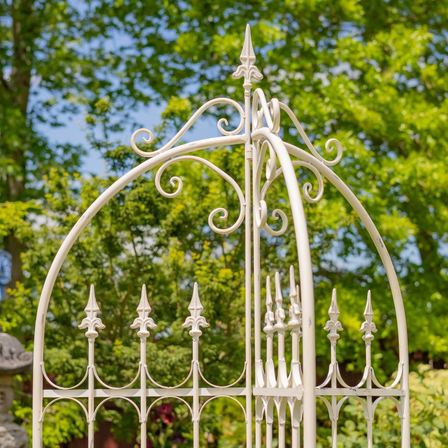 Top of 7.7ft Tall Three Wall Folding Iron Trellis with Fleur-de-lis Accents Paris 1968 with a distressed, hand painted Antique white finish. Image shows a closeup of the top half of the trellis. focusing on the Fleur-de-lis details at the top of the structure, bordered by iron flourish pieces, and again in the trellis section (looking like a metal gate or fence with fleur-de-lis at the top of each horizontal fence