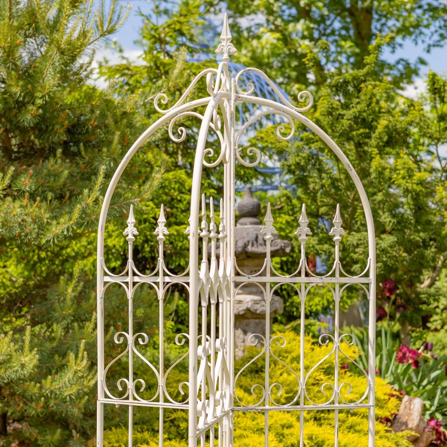 Top of 7.7ft Tall Three Wall Folding Iron Trellis with Fleur-de-lis Accents Paris 1968 with a distressed, hand painted Antique white finish. Image shows a closeup of the top half of the trellis. focusing on the Fleur-de-lis details at the top of the structure, bordered by iron flourish pieces, and again in the trellis section (looking like a metal gate or fence with fleur-de-lis at the top of each horizontal fence