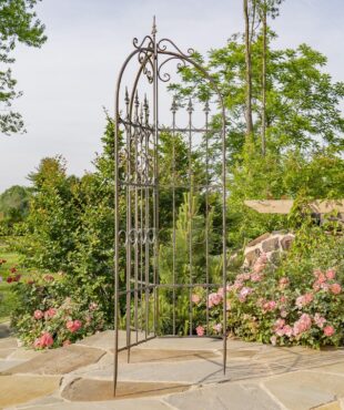 Top of 7.7ft Tall Three Wall Folding Iron Trellis with Fleur-de-lis Accents Paris 1968 with a distressed, hand painted Antique Bronze finish. Image shows the entire outdoor living structure displayed in a garden with beautiful flowers, landscaping and other outdoor décor in the background