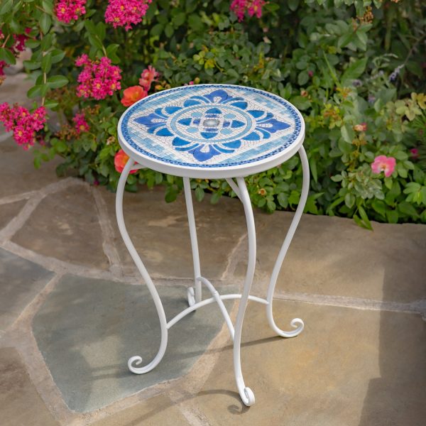 Photo of the Cape May 22in. Tall Mosaic Pedestal Plant Stand on display outdoors on a stone floor with a beautiful blooming rose bush in the background. The plant stand features a glossy white metal frame and White & Blue ceramic tiles with white cement sealing it all together.