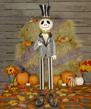 This is a tall Halloween skeleton decoration. He has a big smiling skull, wears a shiny black top hat, a gray jacket with a gold bow tie, and striped pants. He’s holding a stick with a spider web on it. He is standing in front of hay and pumpkins.