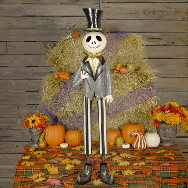 This is a tall Halloween skeleton decoration. He has a big smiling skull, wears a shiny black top hat, a gray jacket with a gold bow tie, and striped pants. He’s holding a stick with a spider web on it. He is standing in front of hay and pumpkins.