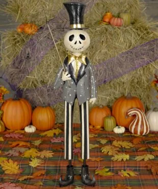 The skeleton is tall and thin, with a round skull head and a stitched smile. It wears a black top hat with a gold band, a gray jacket with white dots, a gold bow tie, and black-and-white striped pants. It holds a stick with a spiderweb and a small black spider hanging from it. The skeleton looks friendly and is dressed like it's ready for a Halloween party.