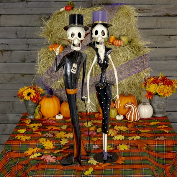 The image shows two tall, thin skeleton figurines dressed in fancy, vintage-style clothing. One is a male skeleton wearing a top hat, black suit with red accents, and holding a cane. The other is a female skeleton in a fitted black dress with white polka dots and a matching top hat. Both have exaggerated features and playful, spooky expressions, like characters from an old horror movie. They are standing side by side against a plain background.